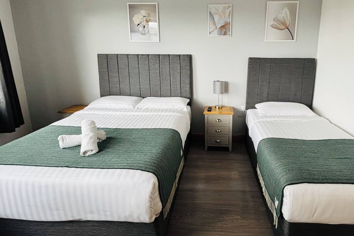 A hotel room with two beds, each with a grey headboard, a green bedspread, and a white pillow. There is a wooden nightstand between the beds with a lamp and a phone.