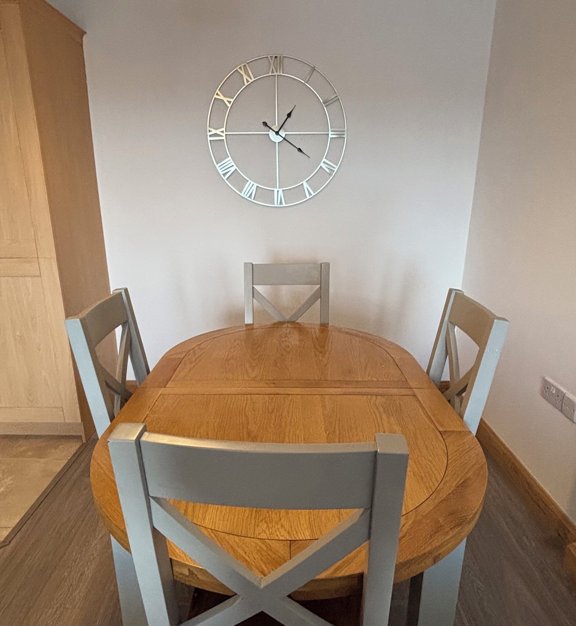 A dining area with a wooden oval table, four light blue chairs, and a wall-mounted clock.