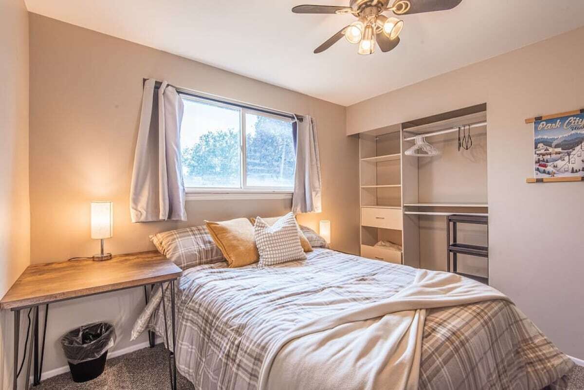 Quarto com cama, mesa de cabeceira, luminária, janela, ventoinha de teto e estantes embutidas.