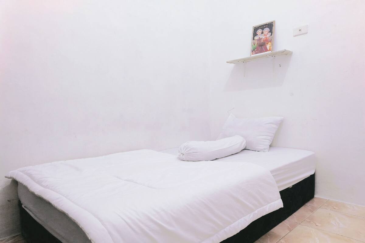 Een kleine kamer met een enkel bed, een witte spreidek en een kussen, plus een klein plankje met een foto.