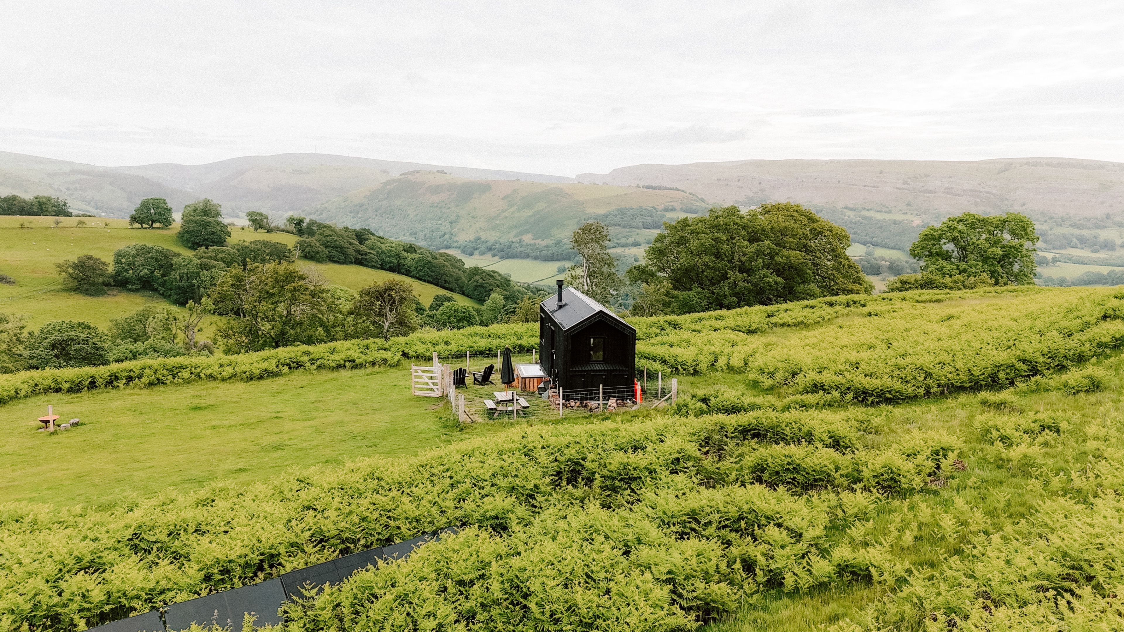 A small cabin with a porch, surrounded by lush greenery and hills.