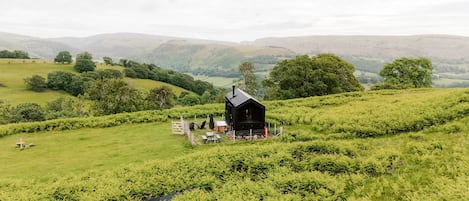 A small cabin with a porch, surrounded by lush greenery and hills.