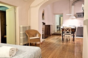 A room with a bed, a wicker chair, a dining table with chairs, a radiator, and a kitchen area with cabinets and a sink.