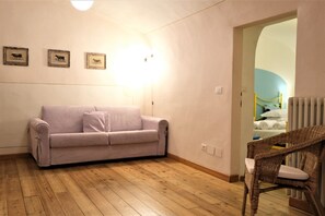 A room with a wooden floor, a light purple sofa, a wicker chair, and a bed with a blue wall in the background.