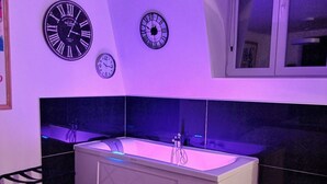 A modern bathroom with a freestanding bathtub, three wall clocks, and a window with blinds. - Les Appartements Fénelon (Le Cateau-Cambresis)