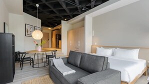 Quarto de hotel moderno com sofá cinzento, cama com roupa de cama branca, área de refeições com mesa redonda e cadeiras, e cozinha ao fundo.