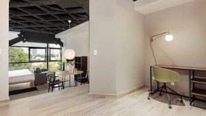 Quarto moderno com uma cama, uma escrivaninha e uma cadeira. Há uma janela com vista para edifícios e árvores.