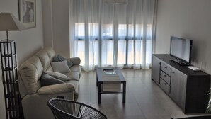 Een moderne woonkamer met een bank, een salontafel, een televisie op een kast en een groot raam met dunne gordijnen.