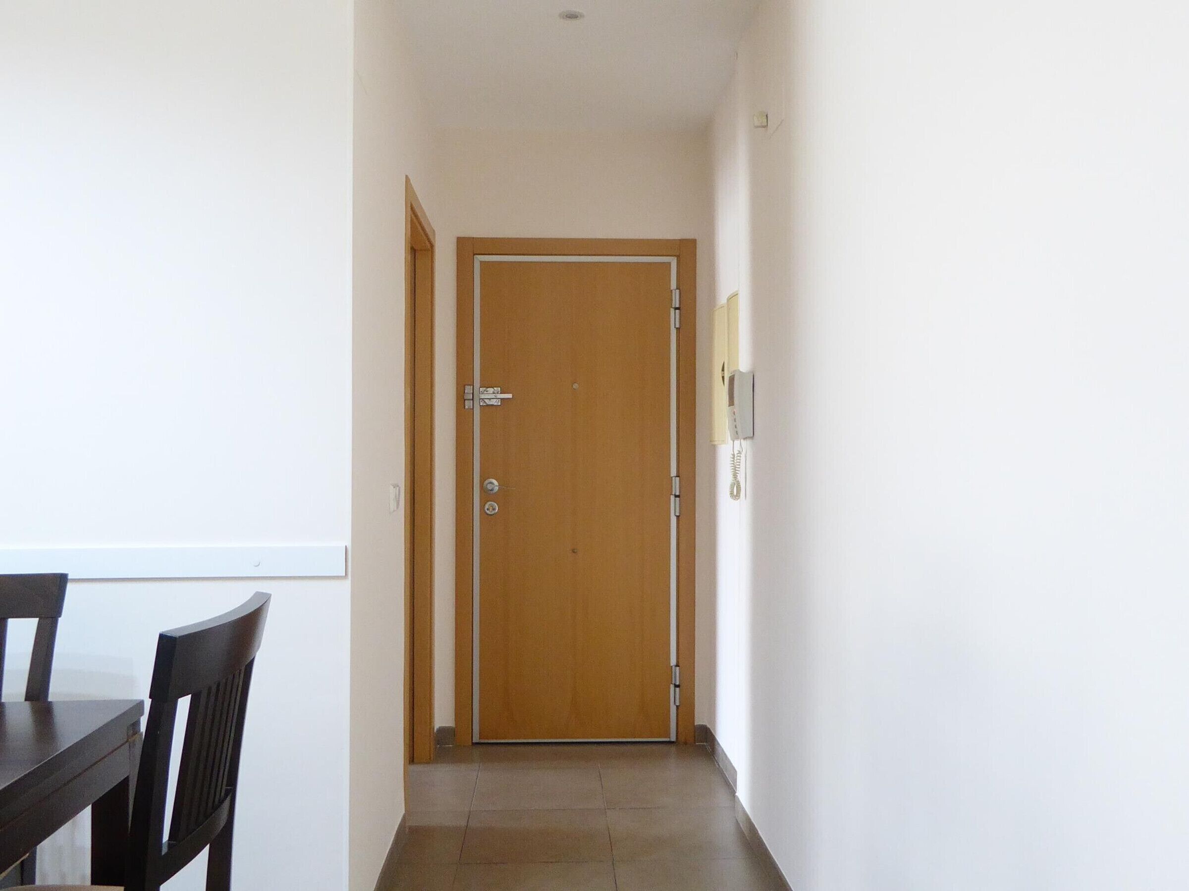 A narrow hallway with a wooden door at the end, a dining table with chairs on the left, and a light switch on the wall to the right.