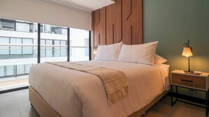 A modern bedroom with a large bed, a wooden headboard, a nightstand with a lamp, and a view of a building through a window. - w* | Captivating 1BR in San Isidro (San Isidro)