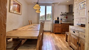 Een houten eettafel met stoelen, een keuken met houten kastjes, een magnetron en een raam met uitzicht op bomen.