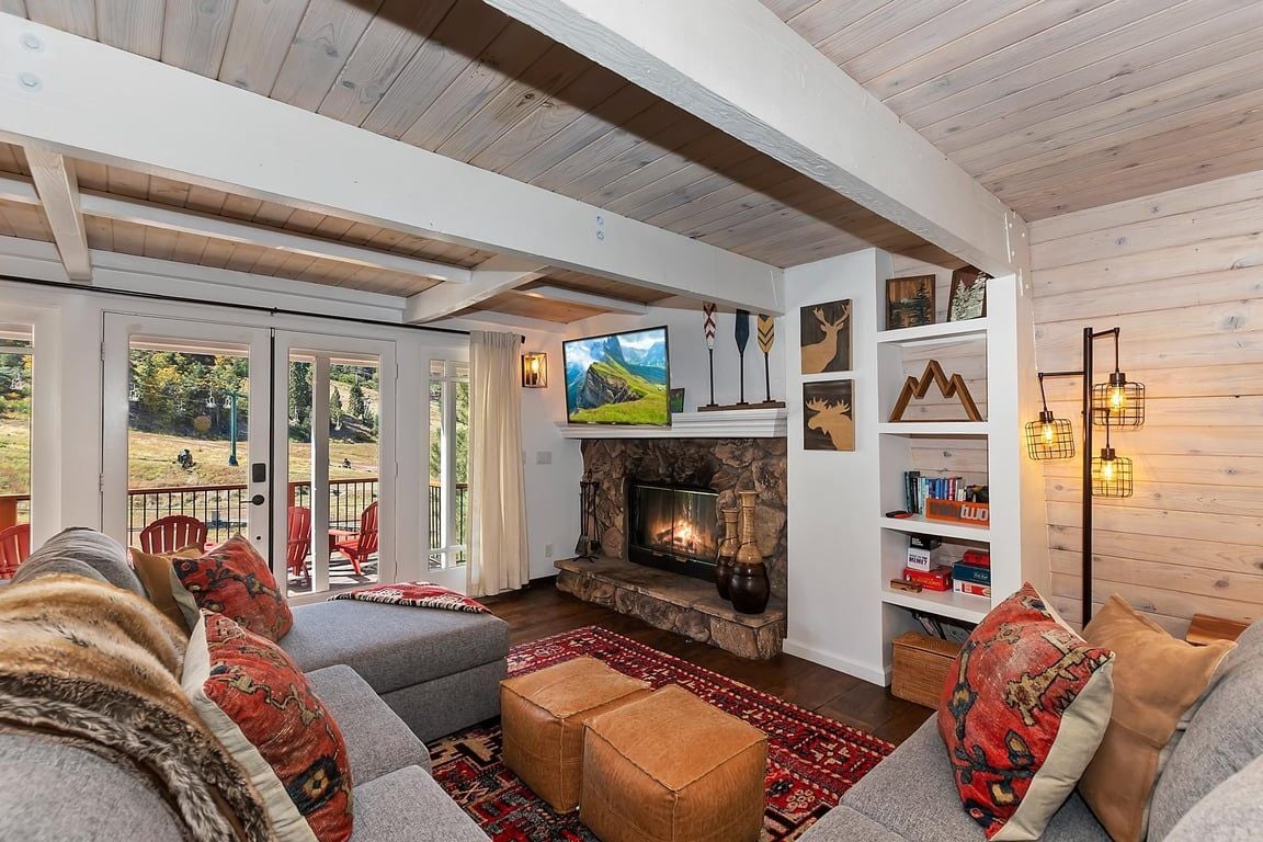 A cozy living room with a fireplace, comfortable seating, and a view through large glass doors.