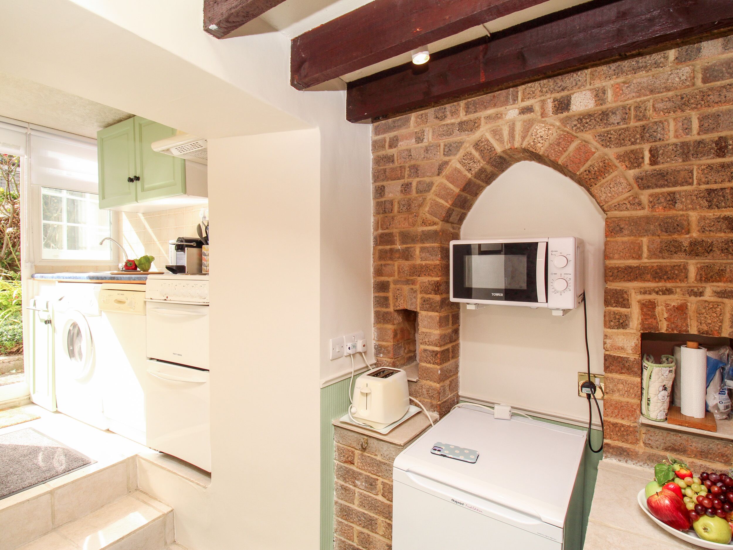 A kitchen with a brick wall, a microwave, a washing machine, and a fruit bowl.
