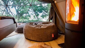 A cozy tent interior with a wood-burning stove, a cushioned seating area, and a view of trees through a geometric window structure. - Luna Domes (Edenbridge)