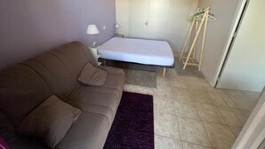 A small, well-lit room with a single bed, a sofa, a small table with a lamp, and a coat rack.