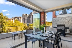 A balcony with a dining table set for two, a grill, and a view of a cityscape. - Mayfair Luxury 212 (Cape Town)