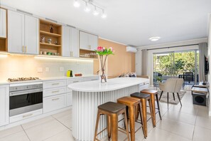 A modern kitchen with white cabinetry, a central island with stools, and a view to an outdoor balcony. - Mayfair Luxury 212 (Cape Town)