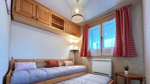 A bedroom with a bed, wooden cabinets, a radiator, a small table with a plant, and a window with curtains. - Charming apartment in Morillon, near cable car (Morillon Village)