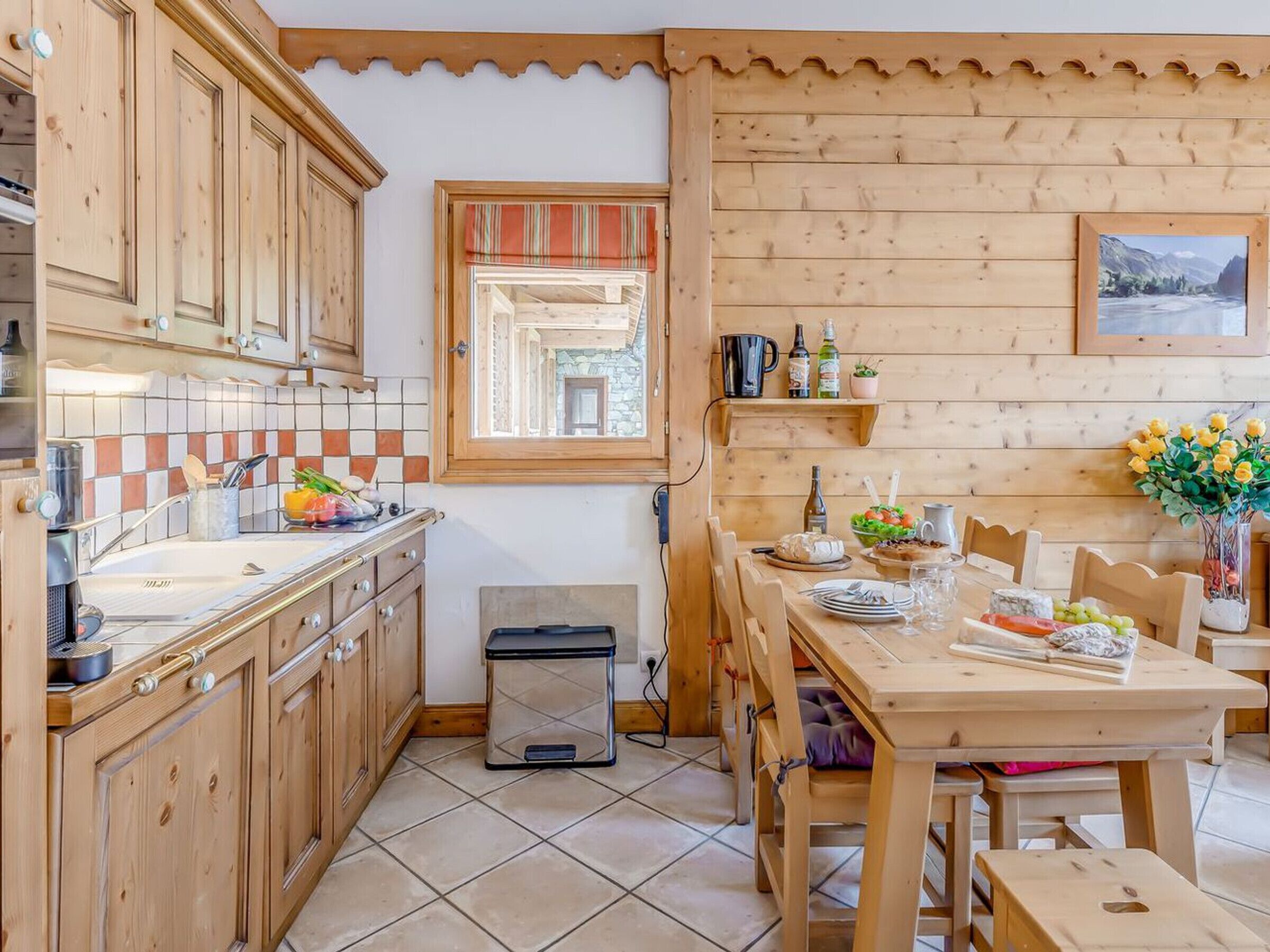 Een keuken met houten kastjes, een betegelde spatwand, een raam met een gestreepte gordijnslag, een eettafel gedekt met borden en glazen, en een kleine prullenbak.