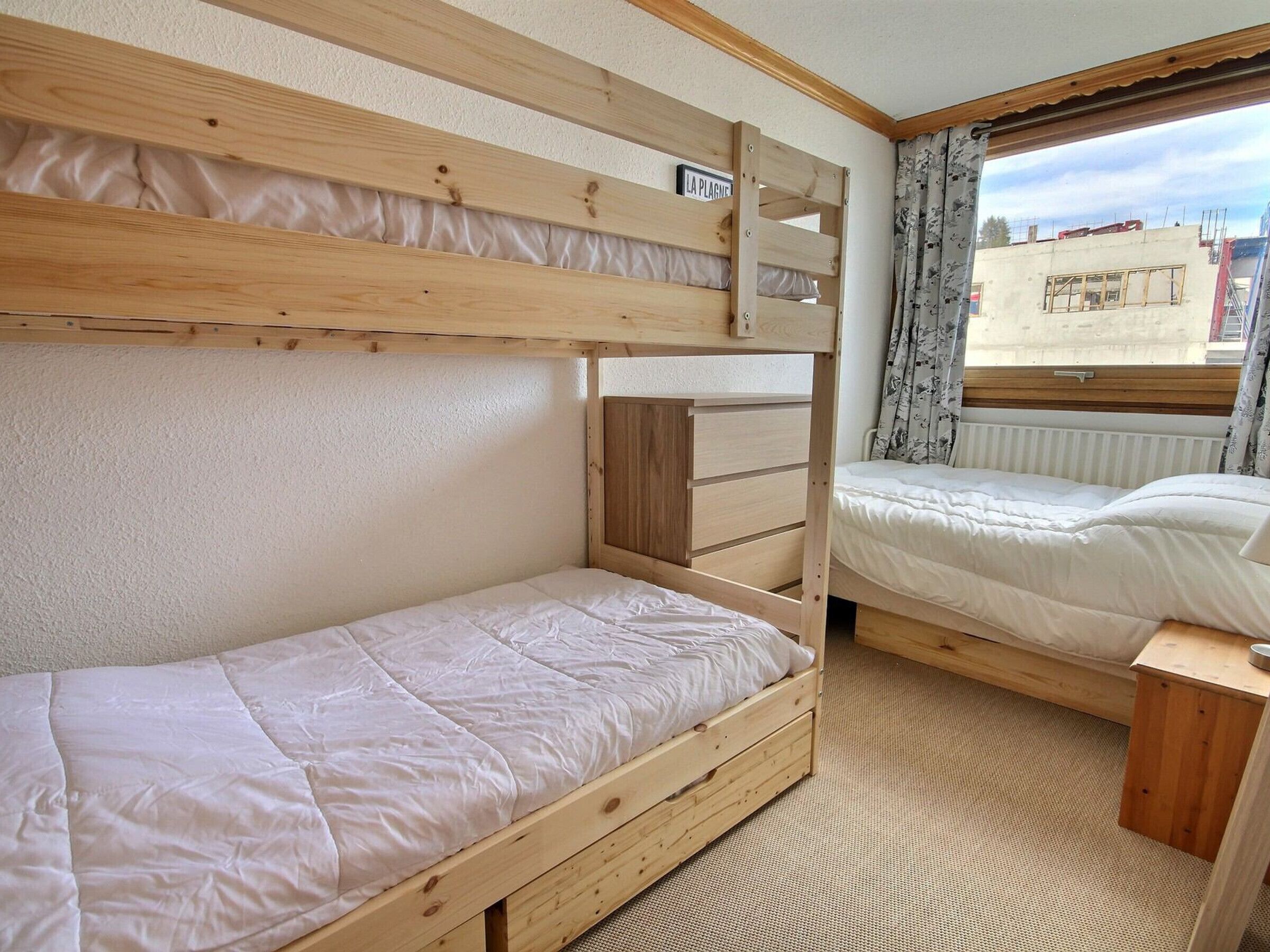 A bunk bed room with a wooden headboard, a small bedside table, and a window with curtains.