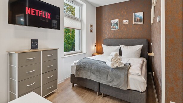 Un dormitorio moderno con una cama, un televisor montado en la pared, una ventana y un aparador.