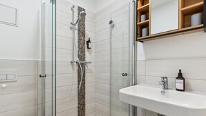 Un baño moderno con ducha envuelta en vidrio, un lavabo blanco con jabón de manos y un estante de madera sobre el lavabo.