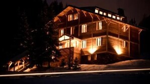 Une cabane en bois à plusieurs étages, avec des fenêtres illuminées, entourée d’arbres et de neige.