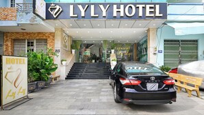 De ingang van Hotel LLY, met een zwarte Toyota geparkeerd ervoor, een trap die naar de ingang leidt en een bord met de hotelnaam.
