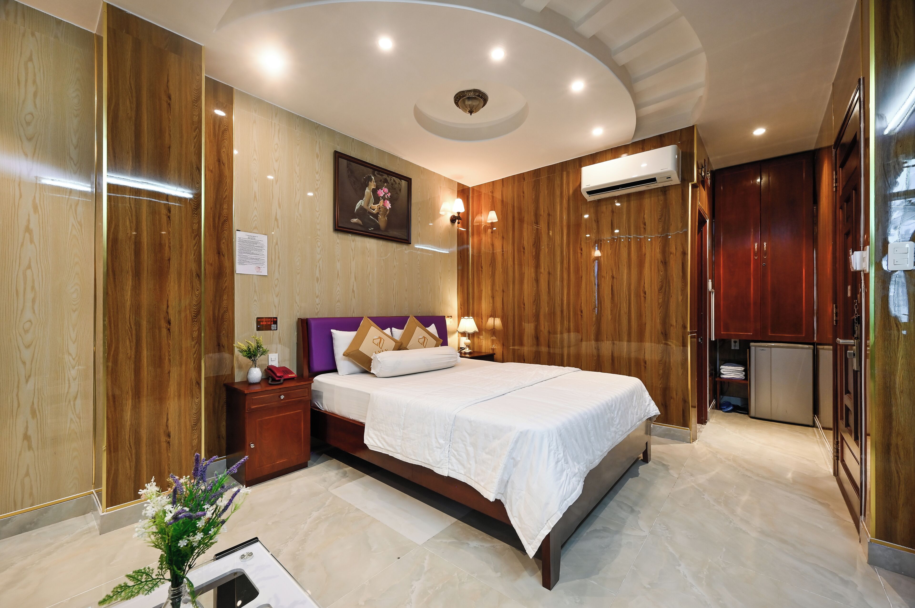 A hotel room with a large bed, wooden paneling, a nightstand, a desk, and a small refrigerator.