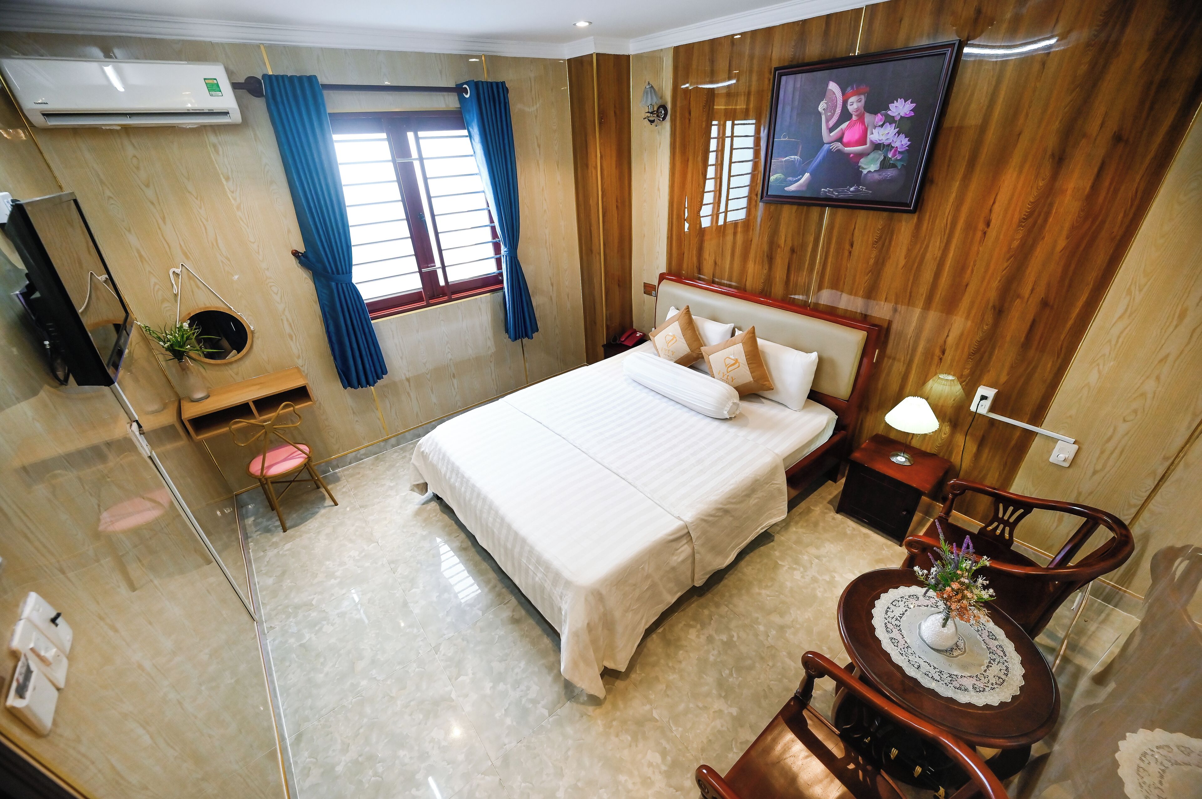 A hotel room with a large bed, a bedside table with a lamp, a wooden wall panel, a window with blue curtains, and a wall-mounted TV.