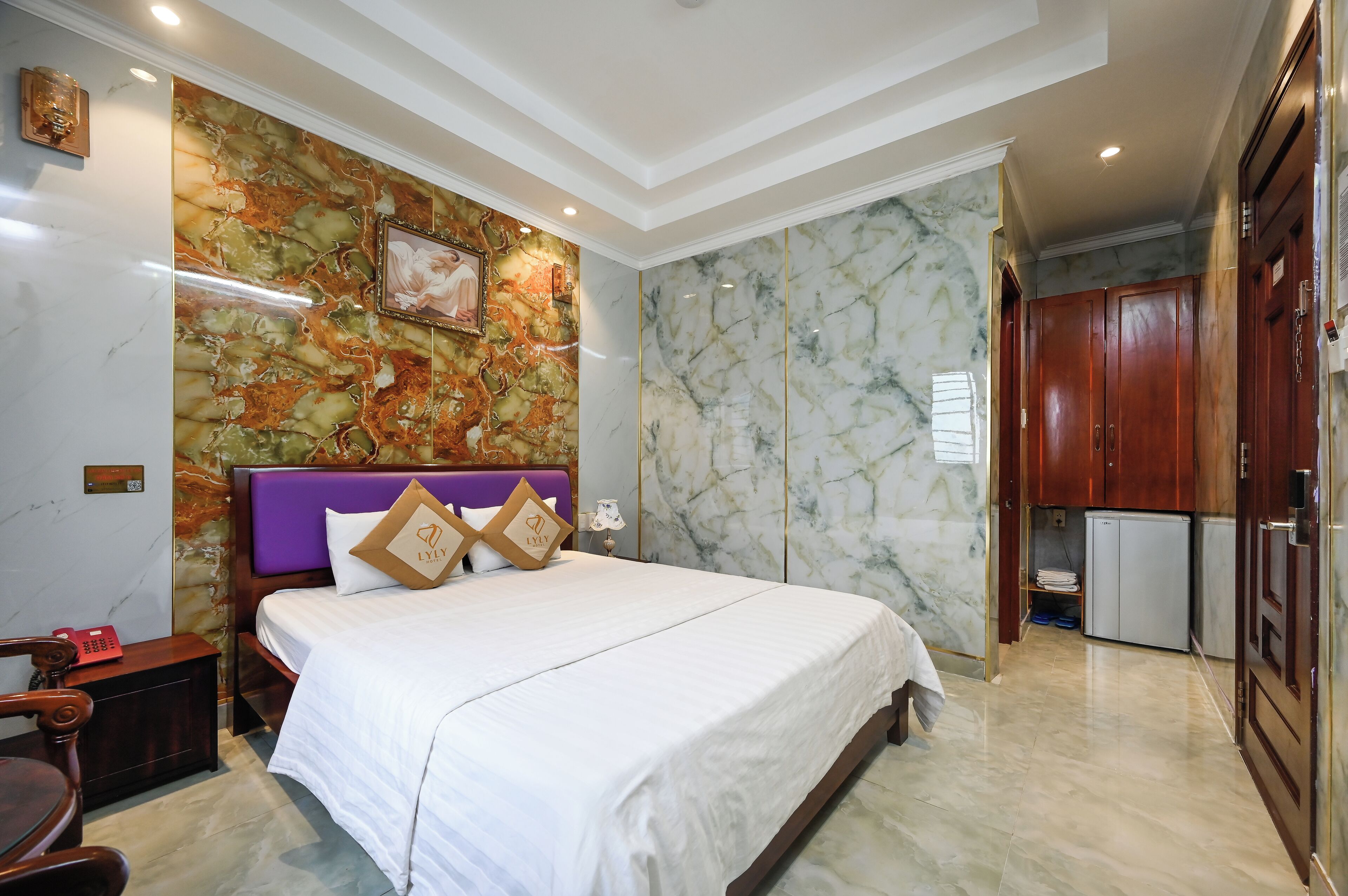A hotel room with a marble-patterned wall, a bed with white bedding, a purple headboard, and a small refrigerator in the corner.