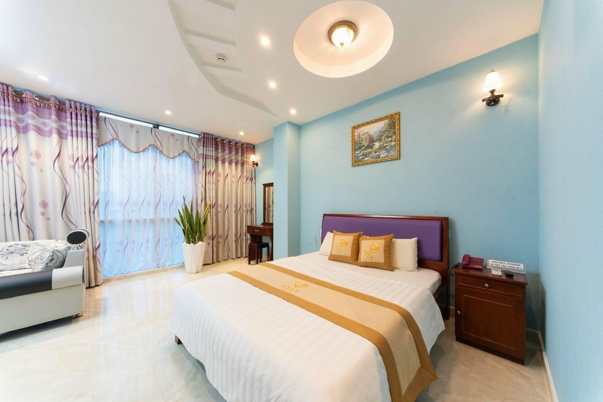 Quarto de hotel com cama, mesa de cabeceira, luminária e uma janela grande com cortinas.