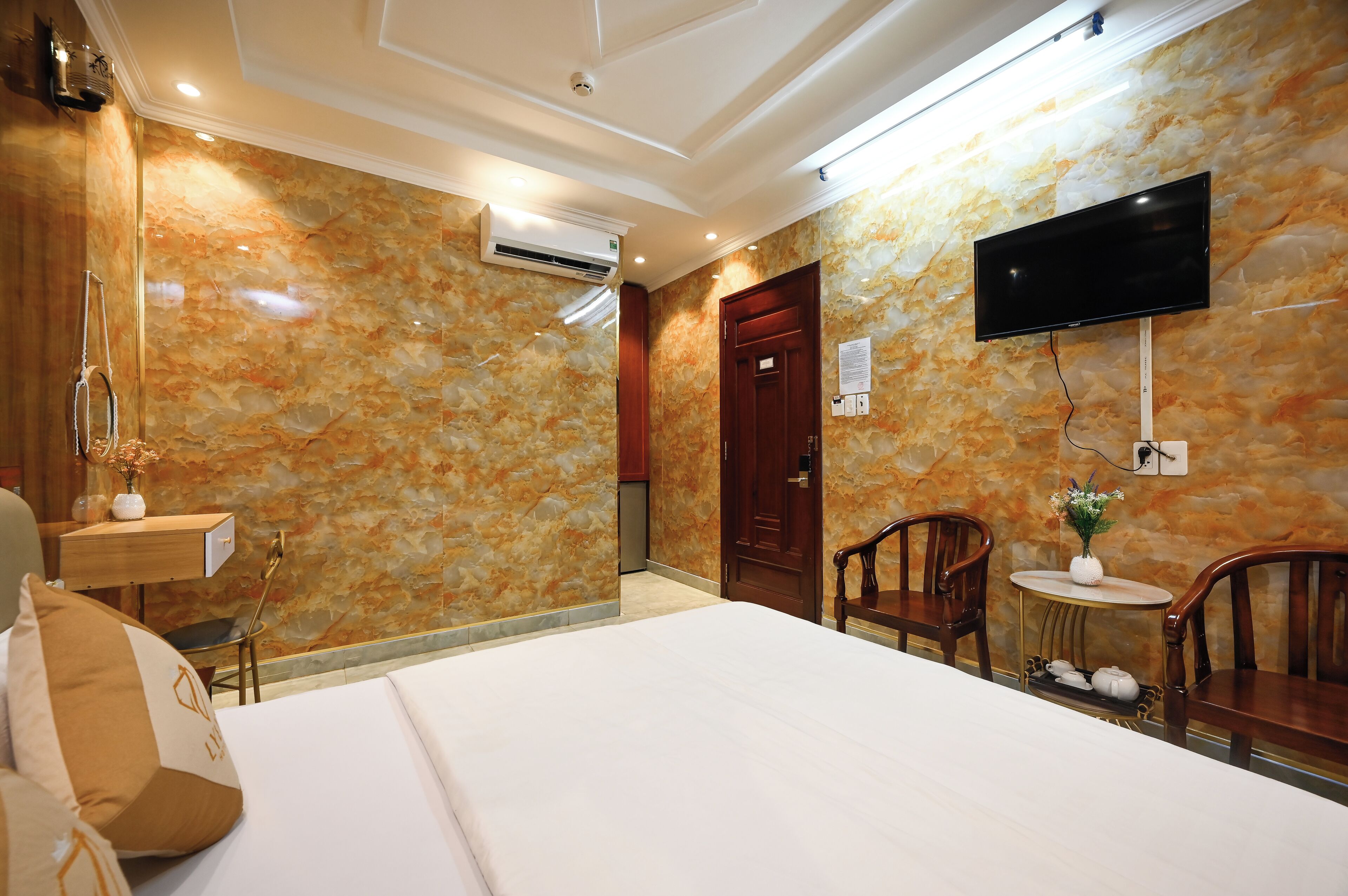 Habitación de hotel con pared de diseño de mármol, una cama, un escritorio con silla, un televisor montado en la pared y una mesita con un jarrón de flores.