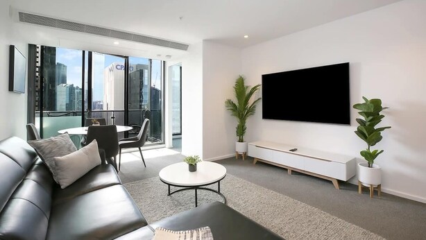 A modern living room with a leather sofa, a round coffee table, and a flat-screen TV mounted on the wall. There are two potted plants and a small table with chairs. - Southbank 2BR Apt City Views, Free Parking (Southbank)