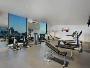 A modern gym with various exercise equipment, large windows offering city views, and a flat-screen TV on the wall.
