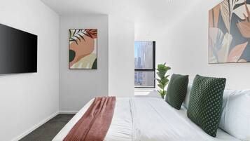 A modern bedroom with a large bed, a flat-screen TV, abstract wall art, and a view of a cityscape through a large window.