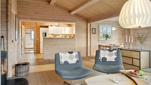 Ein gemütliches Blockhaus-Interieur mit Essbereich, zwei grau gepolsterte Sessel, einem Holztisch, einer Pendelleuchte und Blick auf Küche und Bad durch einen offenen Durchgang.