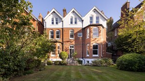 A large, two-story brick house with a well-maintained lawn and a small patio area. - Courtside Charms, by Veeve (London)