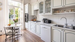 A kitchen with white cabinetry, a dining table with chairs, a window, and a view of greenery outside. - Courtside Charms, by Veeve (London)