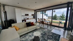 A modern living room with a sofa, coffee table, and dining area. There is a kitchen in the background with cabinets and a countertop. The room has large windows offering a view of the outdoors.