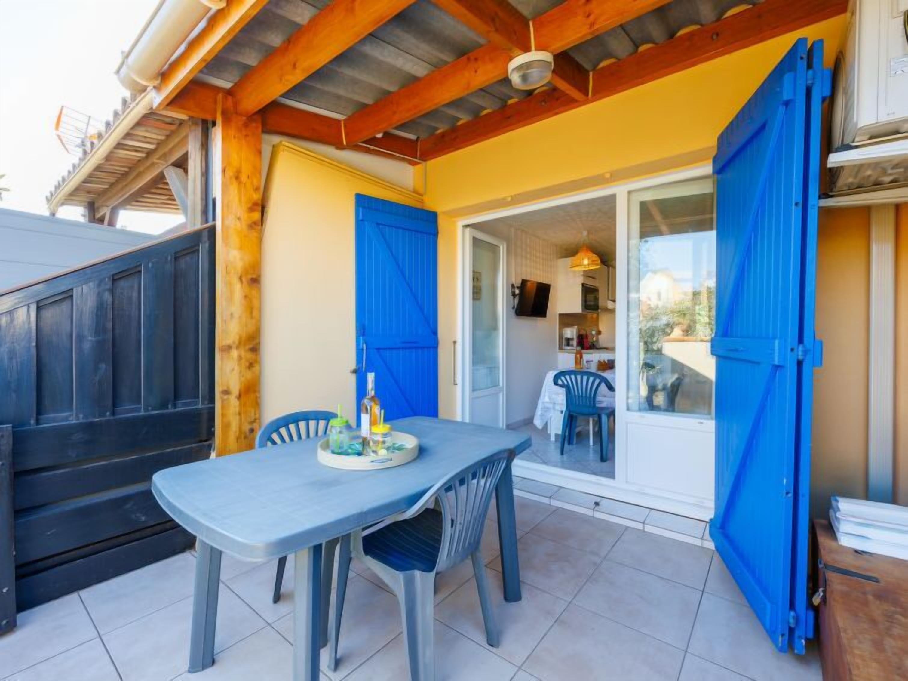 A covered outdoor dining area with a table and chairs, a blue door, and a view into the house.