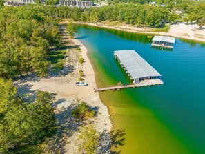 Een luchtbeeld van een landgoed aan een rivier, met een steiger die in het water reikt, een overdekte boathuis, en een wit voertuig geparkeerd op het strand.