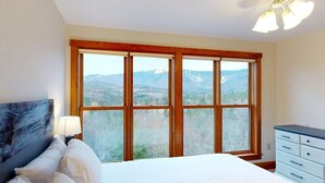A bedroom with a large window offering a mountain view, a bed with white linens, a nightstand with a lamp, and a dresser. - Vermont Post & Beam w/Hot Tub & Stunning Views (Waitsfield)