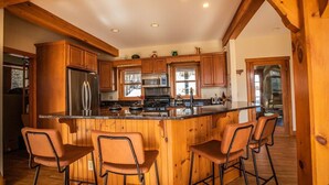 A kitchen with wooden cabinets, a stainless steel refrigerator, a large island with stools, and a view of the outdoors. - Vermont Post & Beam w/Hot Tub & Stunning Views (Waitsfield)