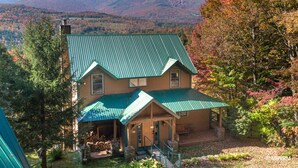 A two-story house with a green metal roof, a covered porch, and a view of mountains in the background. - Vermont Post & Beam w/Hot Tub & Stunning Views (Waitsfield)