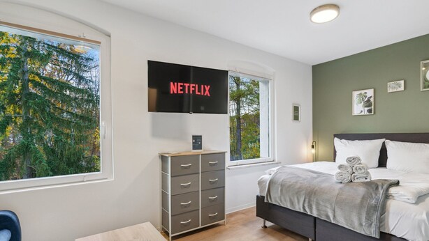 Ein modernes Schlafzimmer mit Bett, Kommode, Wandfernseher und großen Fenstern.