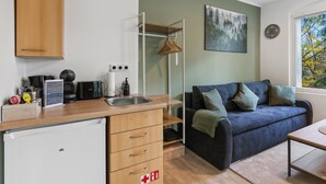 Eine kompakte Küche mit Spüle, Kaffeemaschine und Toaster. Darüber hängt ein Holzschrank, darunter befindet sich ein kleiner Kühlschrank. Eine blaue Couch mit Kissen lehnt an der Wand, darüber hängt ein Gemälde.