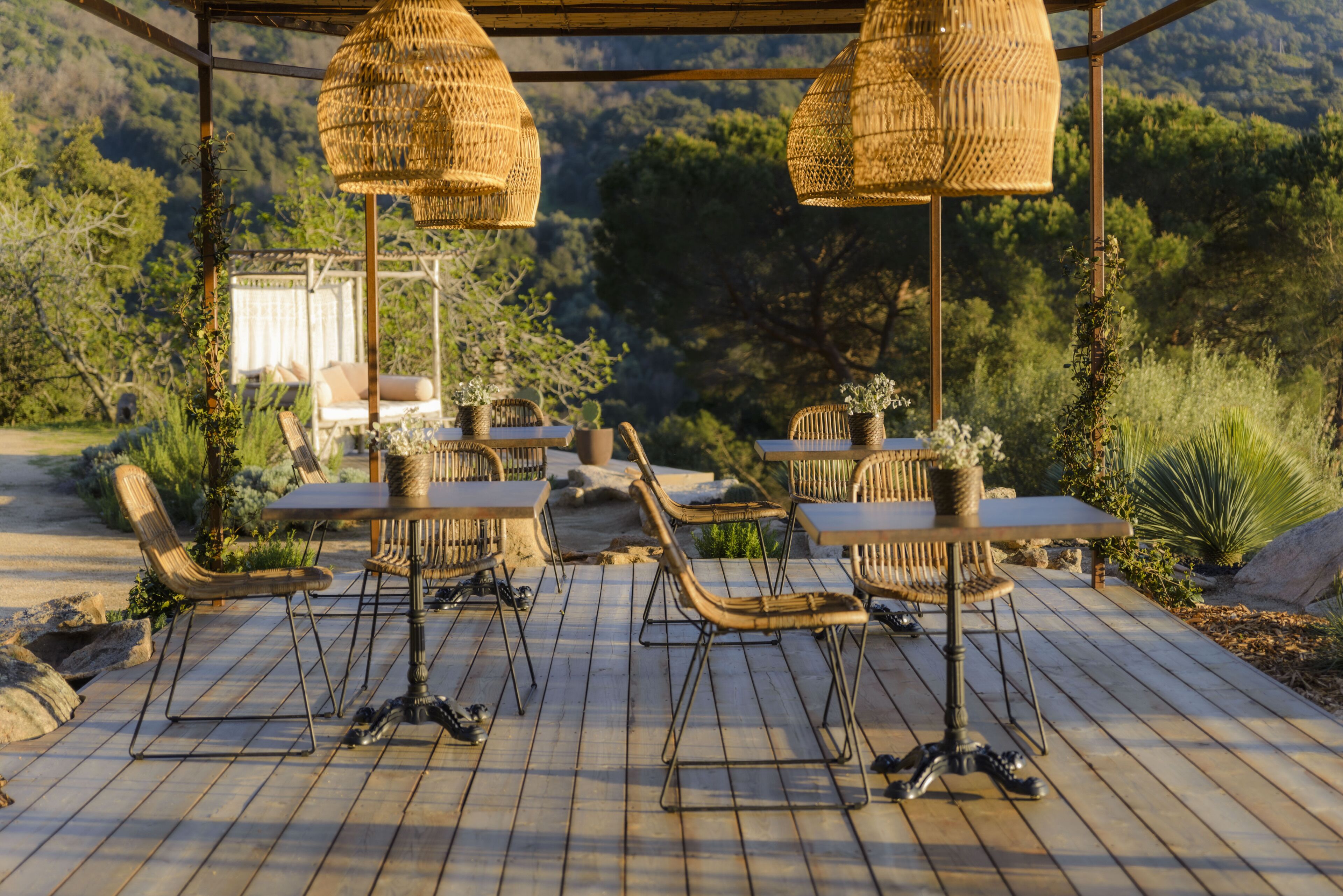 Patio esterno con mobili in vimini e luci pendenti, con sullo sfondo vegetazione rigogliosa e colline.