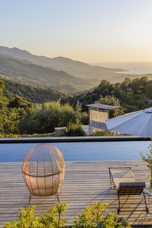 Une terrasse en bois avec une piscine, un fauteuil en osier et une chaise longue donnant sur un paysage montagneux.