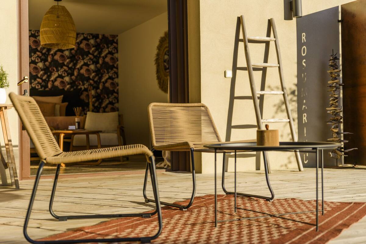 Un espace patio comprenant deux fauteuils en osier, une petite table, une échelle et un mur décoratif portant l’inscription « ROS MARIA ».